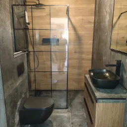 Nowoczesna łazienka z kabiną prysznicową typu walk-in z czarnymi profilami, wiszącą toaletą w kolorze antracytowym oraz umywalką nablatową na drewnianej szafce z szarym blatem.