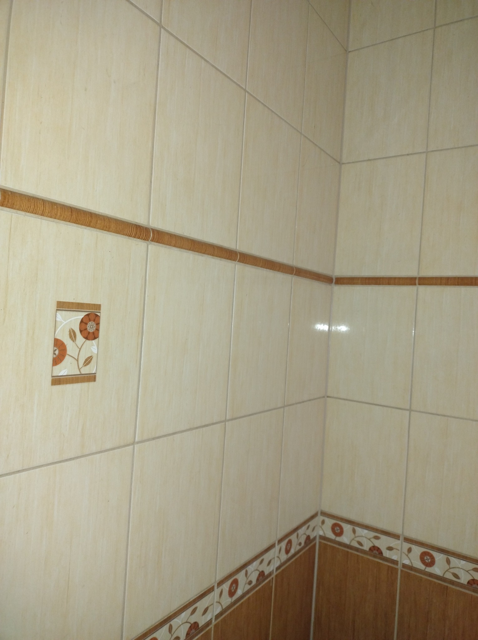 Beżowe płytki ścienne w łazience z brązowym dekoracyjnym pasem z motywem kwiatowym i pojedynczym kwadratowym elementem dekoracyjnym z kwiatami.