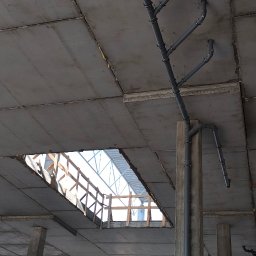kosikredkipl - Szare rury kanalizacyjne zamontowane pod betonowym stropem w surowym stanie, widoczne mocowania i elementy instalacji. W tle otwór w stropie i elementy konstrukcyjne.