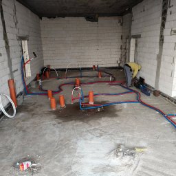 kosikredkipl - Instalacja wodna i kanalizacyjna w surowym wnętrzu. Czerwono-niebieskie rury ułożone na podłodze, pomarańczowe piony kanalizacyjne. Hydraulik w trakcie pracy, poziomica obok.
