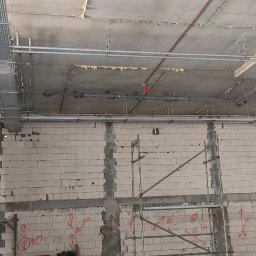 kosikredkipl - Surowy stan: instalacje pod stropem, rury i koryta kablowe w nowym budynku. Ściany z betonu komórkowego z zaznaczeniami. Widoczne rusztowania.