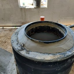 kosikredkipl - Betonowy zbiornik z czarnym pierścieniem i świeżym cementem, widoczny w surowym wnętrzu budynku w trakcie budowy. Pomarańczowa rura kanalizacyjna z boku.