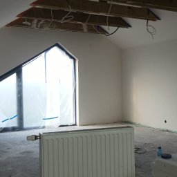 Pro bud - Poddasze w trakcie remontu: okno zabezpieczone folią, grzejnik, surowy beton na podłodze, odsłonięta konstrukcja dachu i instalacje na suficie, ściany przygotowane do malowania.