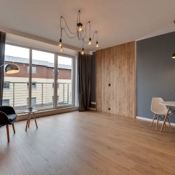 Pro bud - Jasne, przestronne wnętrze z drewnianą podłogą, szarą ścianą, zabudową z paneli imitujących drewno, dużym oknem z szarymi zasłonami, lampą podłogową, krzesłem z narzutą, stolikiem kawowym...