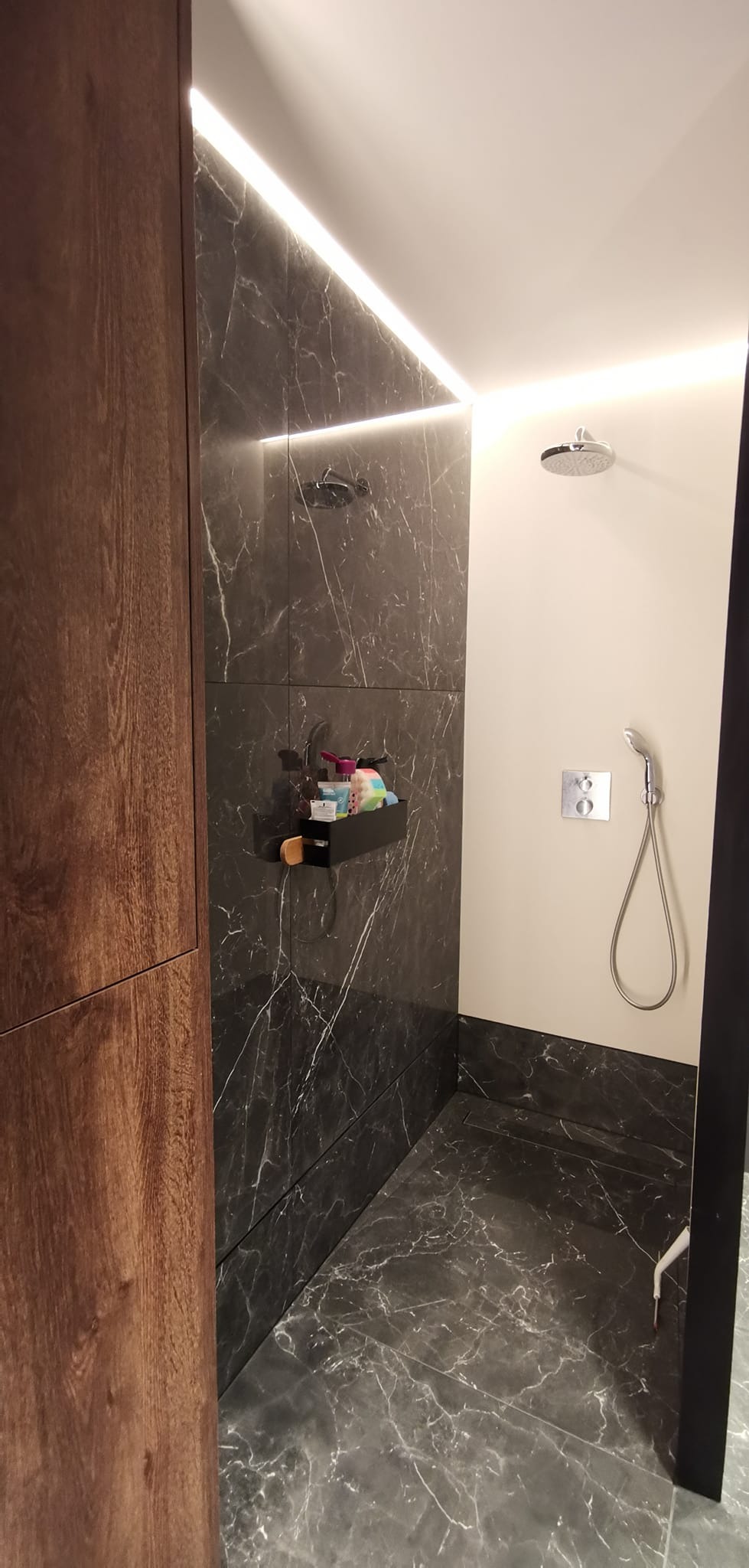 Nowoczesna kabina prysznicowa z marmurowymi płytkami, podświetleniem LED i drewnianą szafką, wyposażona w czarną półkę z kosmetykami i odpływ liniowy.