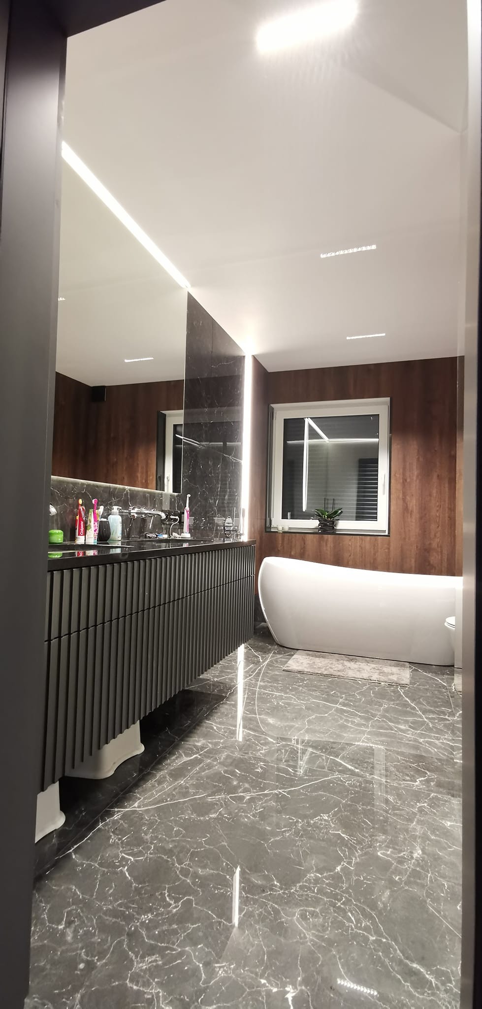 Nowoczesna łazienka z szarymi marmurowymi płytkami podłogowymi, czarną szafką umywalkową z pionowymi żłobieniami, wolnostojącą białą wanną i oknem z roletami.