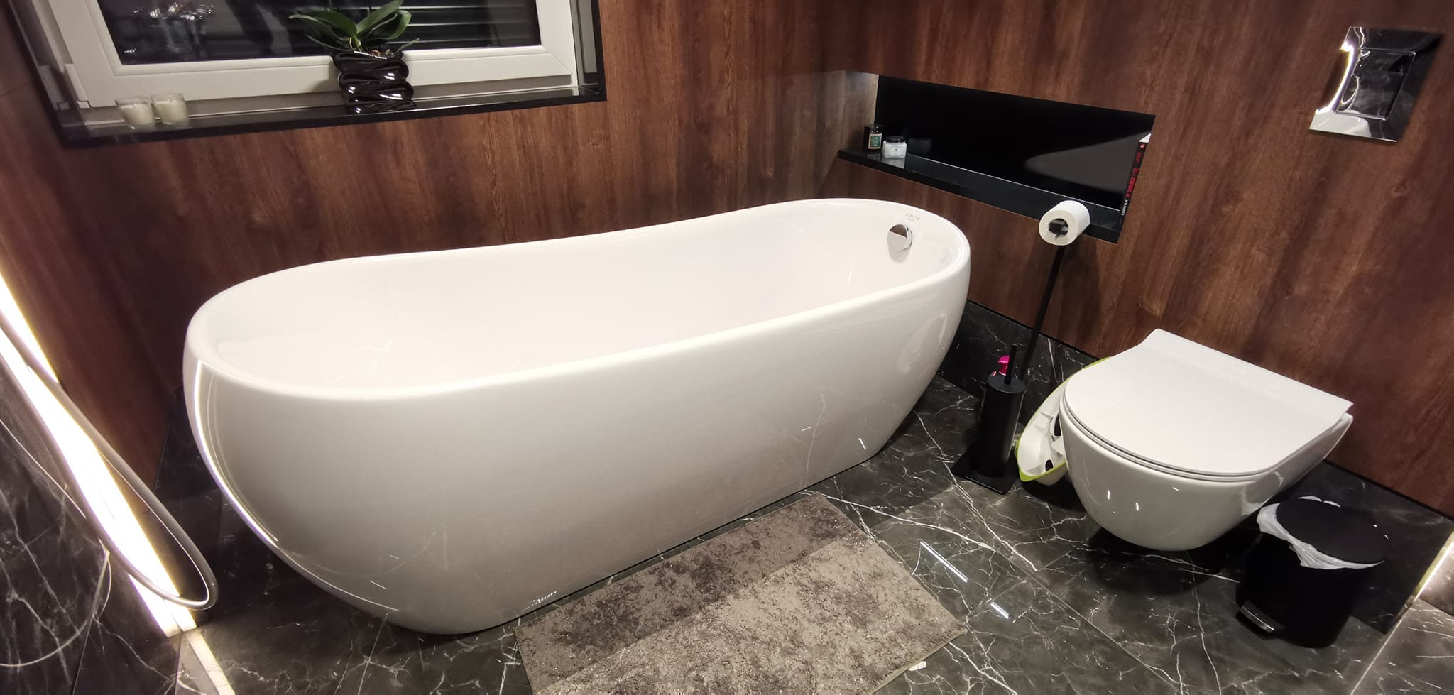 Nowoczesna łazienka z wolnostojącą białą wanną, podwieszaną toaletą, drewnopodobnymi panelami ściennymi i marmurową podłogą, uzupełniona czarnymi akcentami w postaci półki, szczotki do WC i kosza...