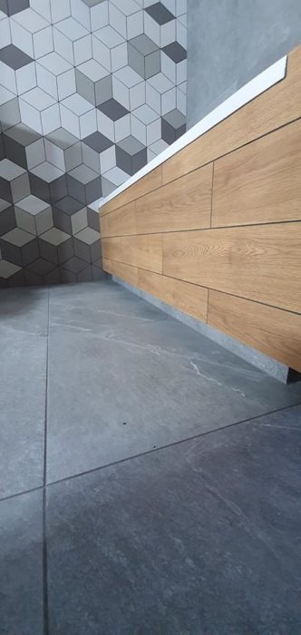 Nowoczesna łazienka z drewnianą zabudową umywalki, szarą podłogą z płytek imitujących kamień i ścianą wykończoną geometrycznymi płytkami w odcieniach szarości i bieli.