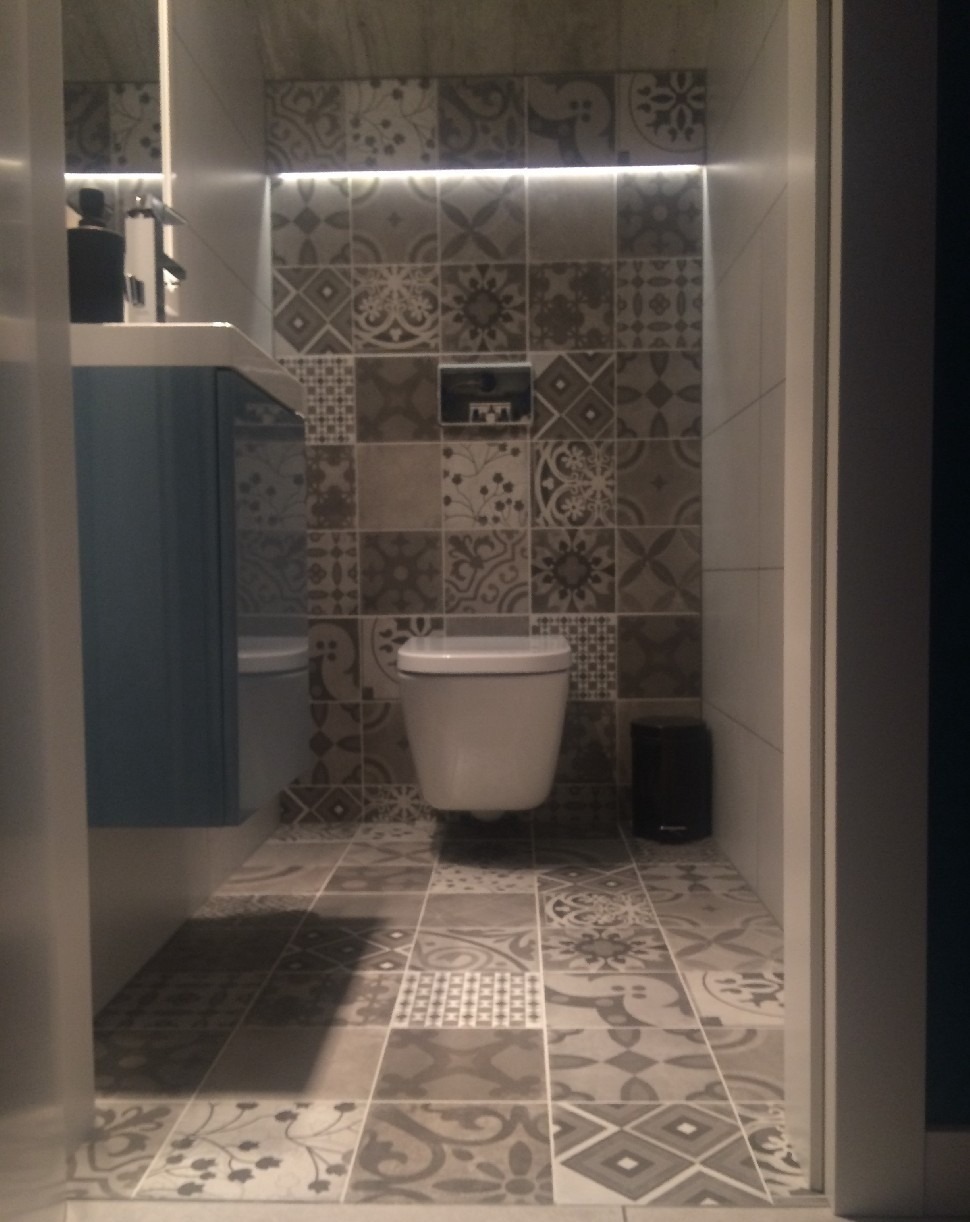 Wykończona łazienka w Skierniewicach z wzorzystymi płytkami na ścianach i podłodze, podświetleniem LED nad toaletą i szafką umywalkową w odcieniach niebieskiego.