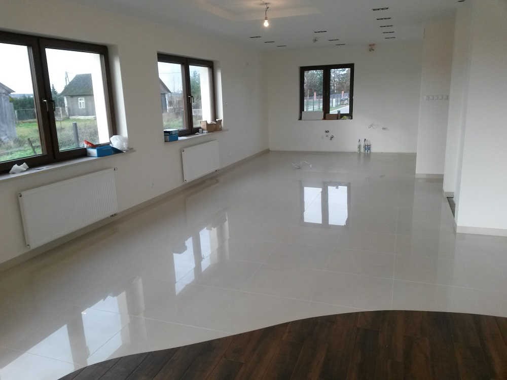 Jasne, błyszczące płytki podłogowe w pustym pokoju z trzema oknami i białymi ścianami; widoczne grzejniki pod oknami i przygotowane punkty instalacyjne na ścianie.