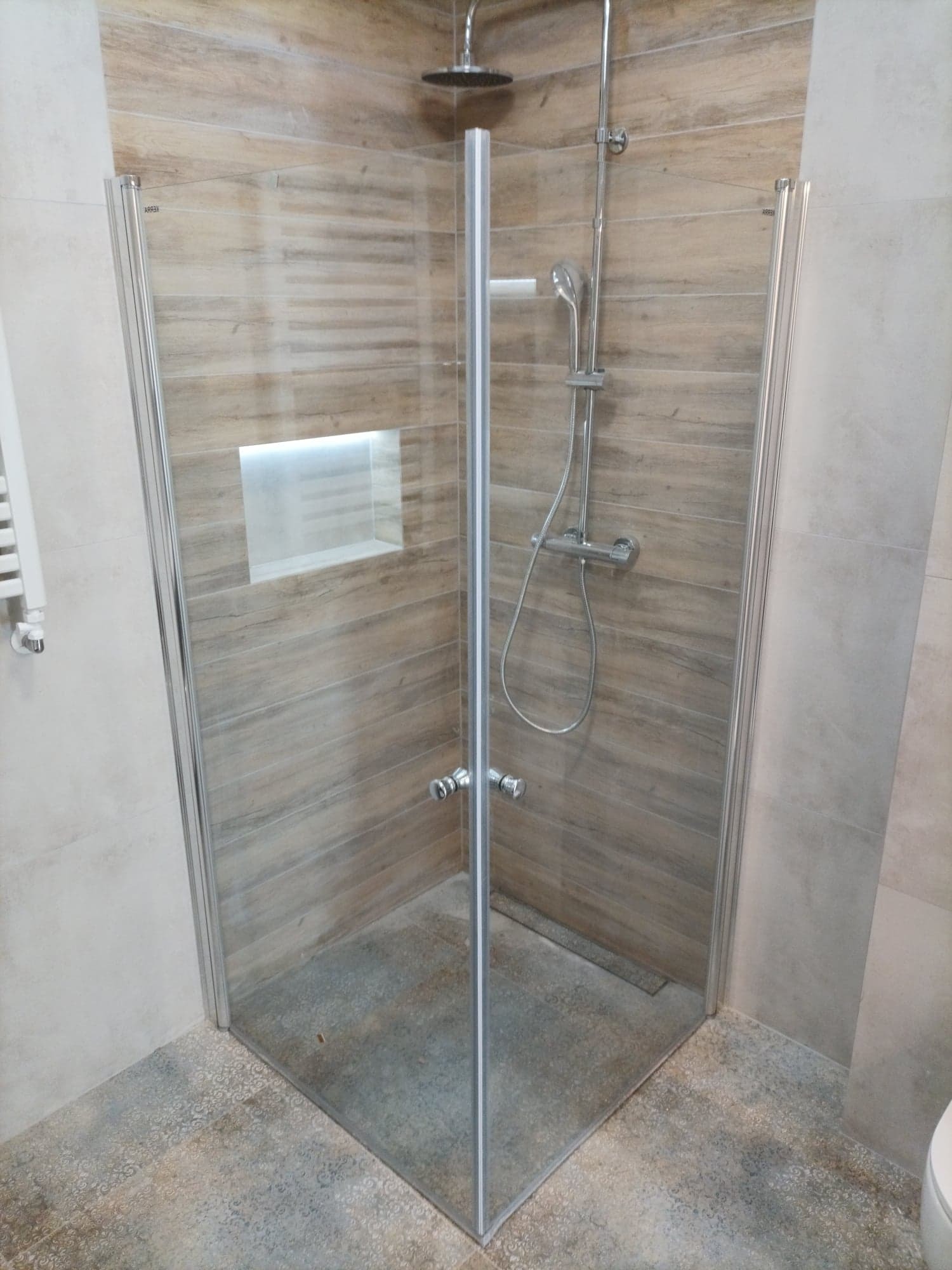 Nowoczesna kabina prysznicowa z chromowanymi detalami, szklanymi drzwiami i płytkami imitującymi drewno na ścianach, wbudowana półka z oświetleniem, widok z zewnątrz.