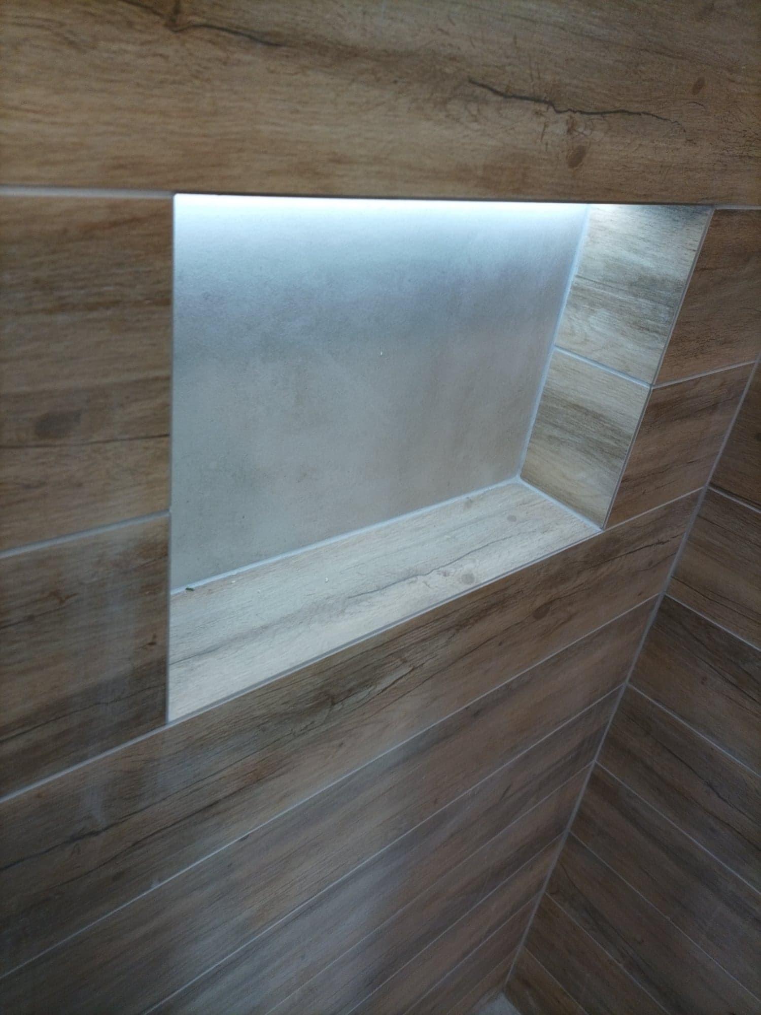 Płytki imitujące drewno ułożone w jodełkę na ścianie i podłodze łazienki, z podświetloną wnęką wykończoną tym samym materiałem.