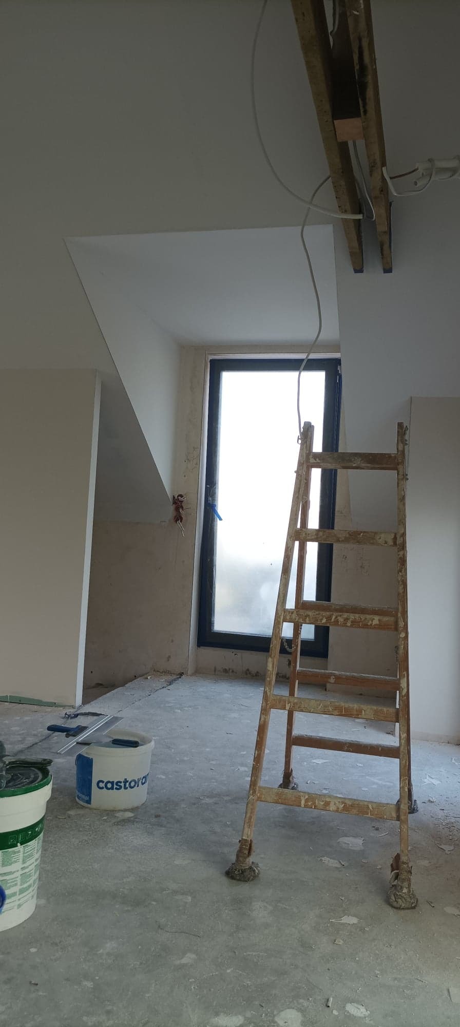 Wnętrze w trakcie prac wykończeniowych: drewniana drabina oparta o ścianę z oknem, widoczne wiadra z farbą i narzędzia, surowe ściany przygotowane do malowania.