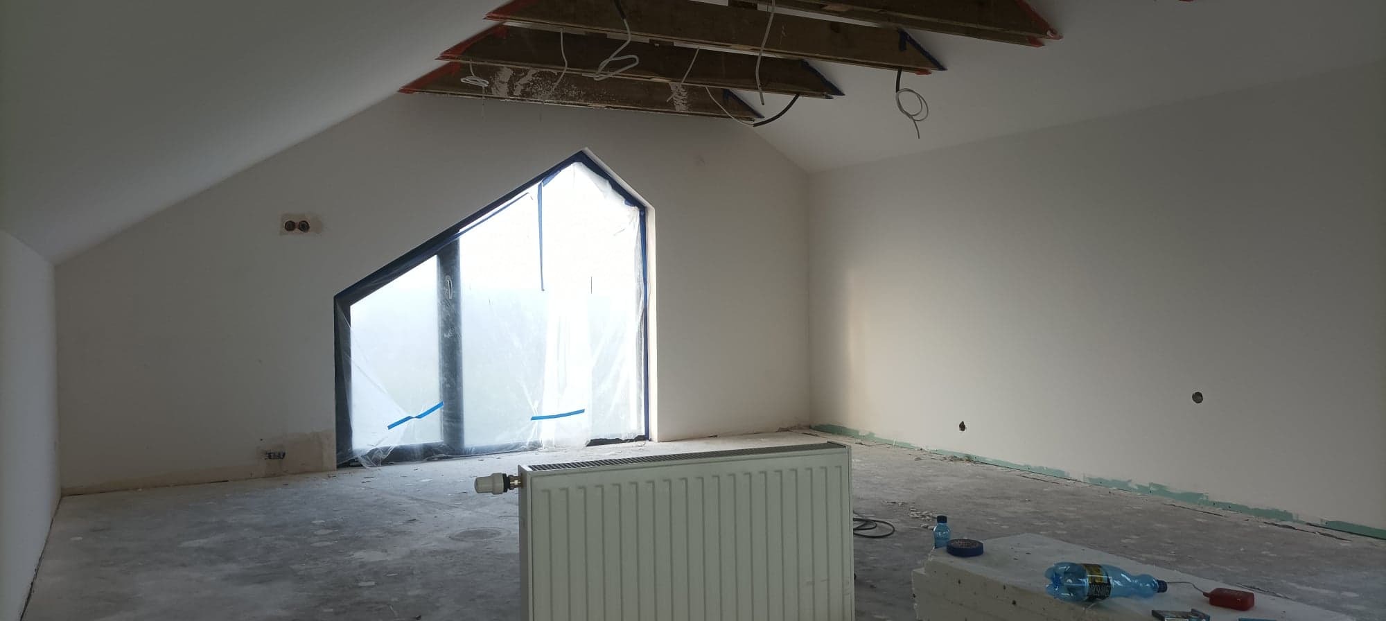 Poddasze w trakcie remontu: okno zabezpieczone folią, grzejnik, surowy beton na podłodze, odsłonięta konstrukcja dachu i instalacje na suficie, ściany przygotowane do malowania.