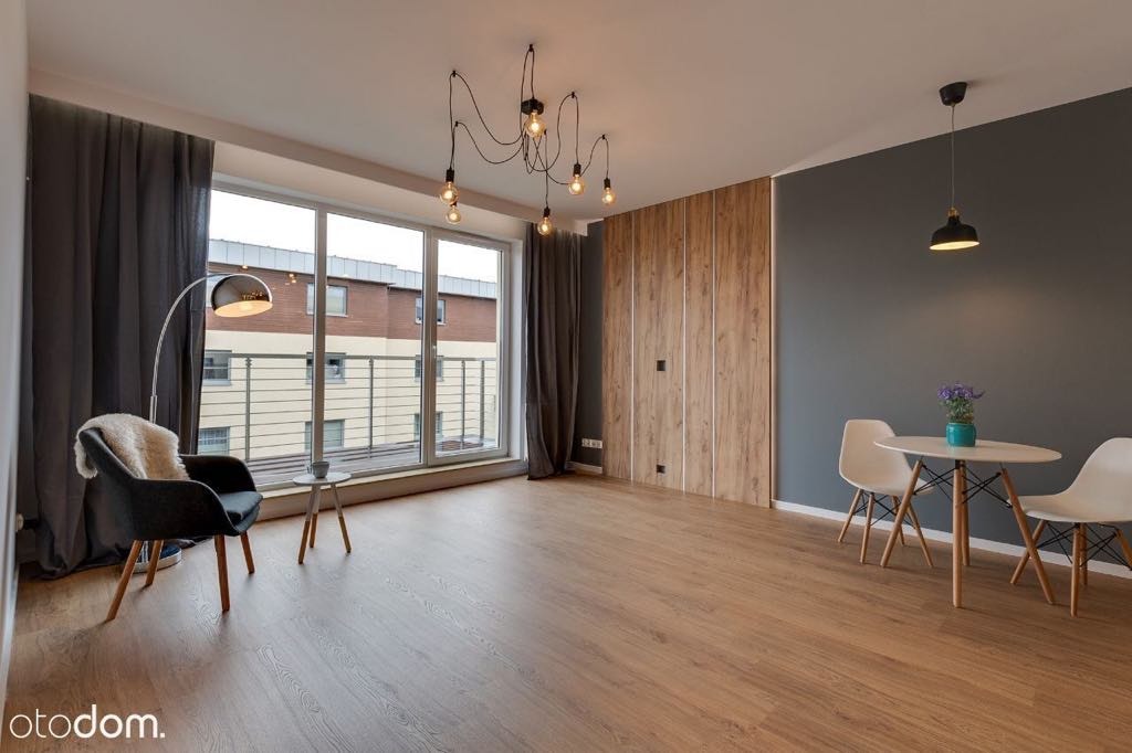 Jasne, przestronne wnętrze z drewnianą podłogą, szarą ścianą, zabudową z paneli imitujących drewno, dużym oknem z szarymi zasłonami, lampą podłogową, krzesłem z narzutą, stolikiem kawowym...