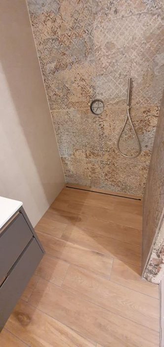 Wykończona kabina prysznicowa z imitacją drewnianej podłogi i wzorzystymi płytkami ściennymi w odcieniach beżu i brązu, widoczny odpływ liniowy i bateria prysznicowa.