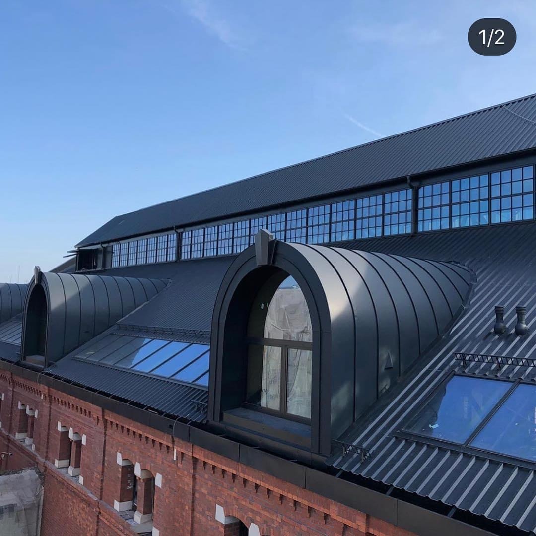 Czarny dach z blachy trapezowej z charakterystycznymi lukarnami w kształcie łuku na ceglanym budynku, widok z góry przy słonecznej pogodzie.
