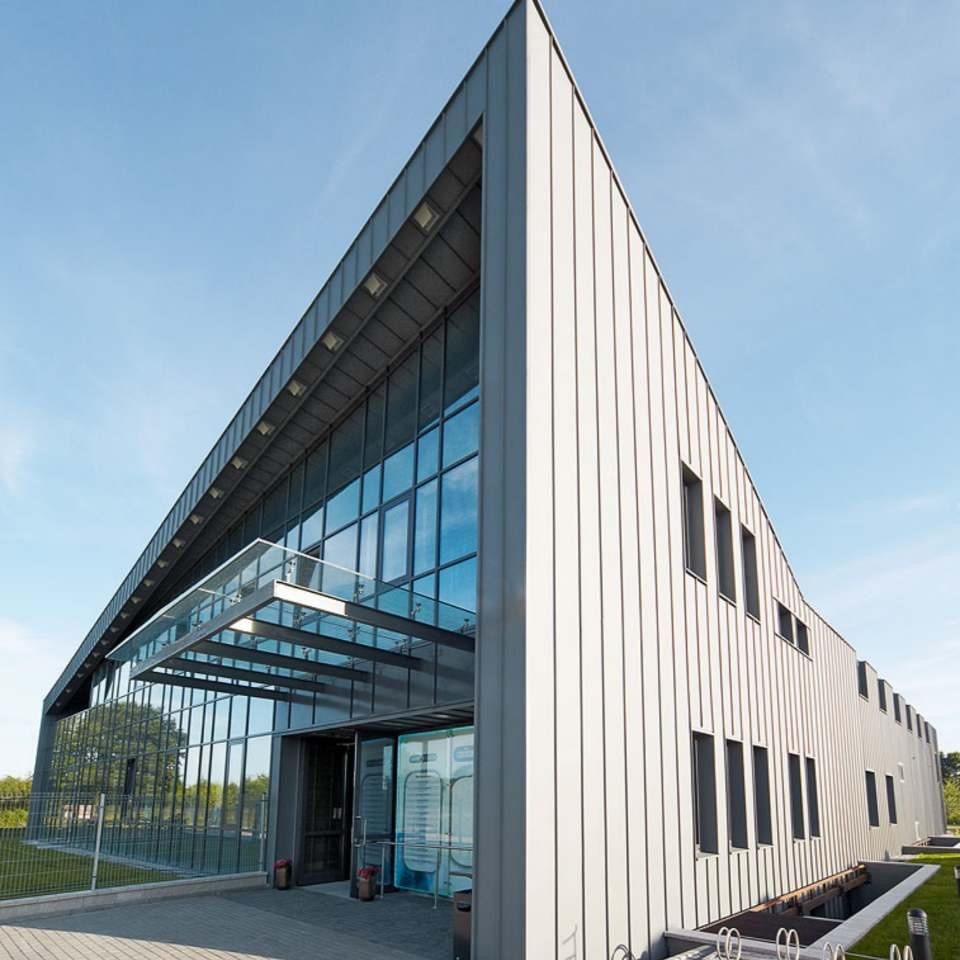 Nowoczesny budynek biurowy z elewacją z paneli pionowych w odcieniach szarości, przeszklonym wejściem i zadaszeniem, widziany z perspektywy żabiej z błękitnym niebem w tle.