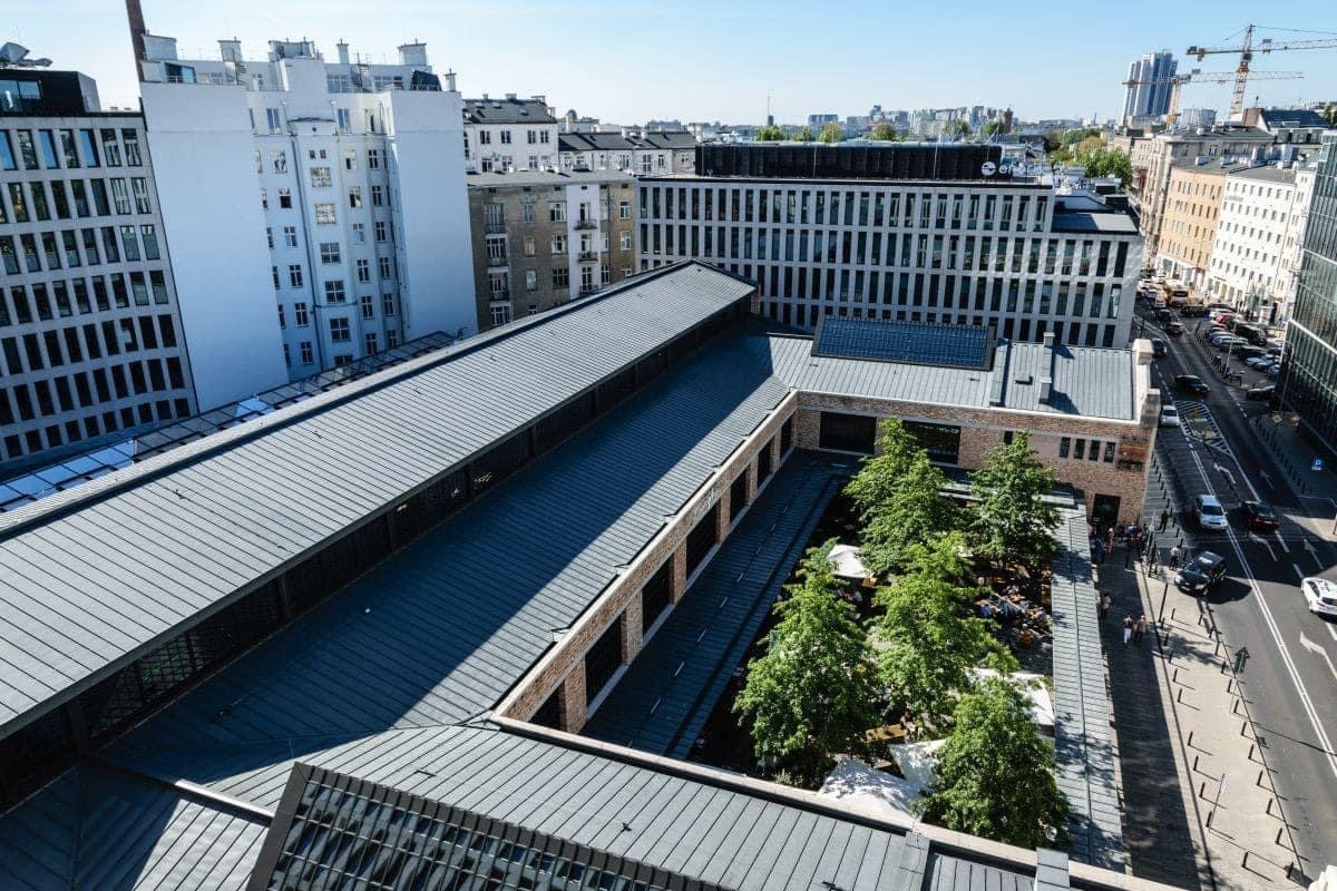 Widok z góry na budynek z szarą blachą trapezową na dachu, z wewnętrznym dziedzińcem z drzewami, otoczony innymi budynkami i ulicą z samochodami w słoneczny dzień.