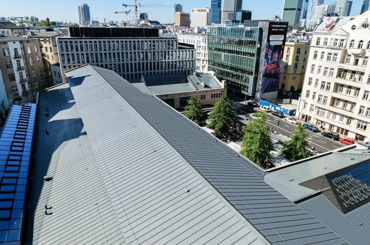 Widok z góry na duży, płaski dach z blachy w kolorze szarym, z widocznymi liniami łączeń, na tle miejskiego krajobrazu z wysokimi budynkami i zielenią drzew.