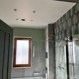 Wykończona łazienka z wanną, przeszklonym prysznicem walk-in, drewnianym oknem i mozaiką z pionowych płytek w odcieniach szarości i błękitu, nowoczesne oświetlenie sufitowe i ścienne.