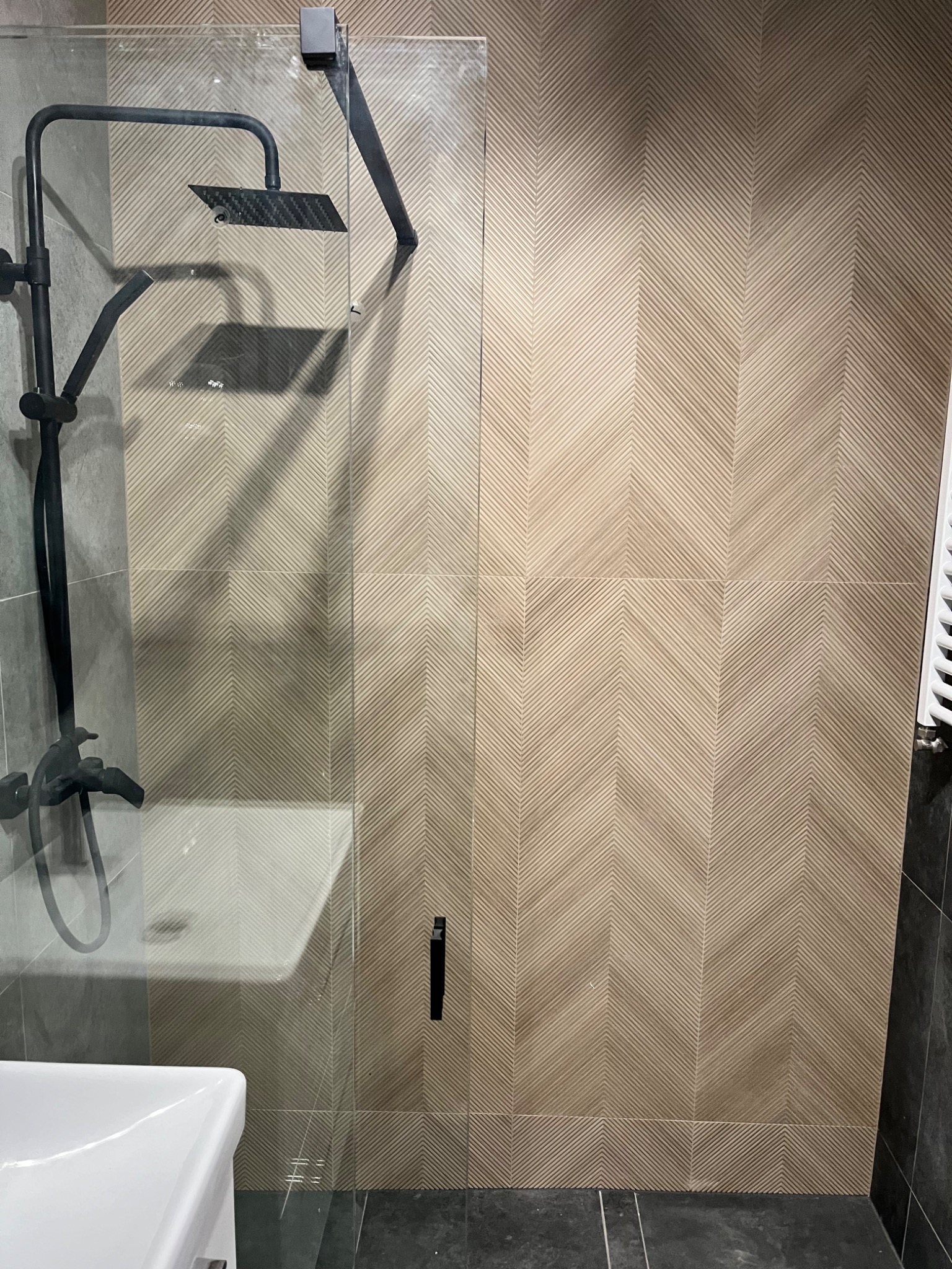 Nowoczesna kabina prysznicowa z czarną armaturą i deszczownicą, widoczna umywalka, ściana wyłożona płytkami w jodełkę w odcieniach beżu.
