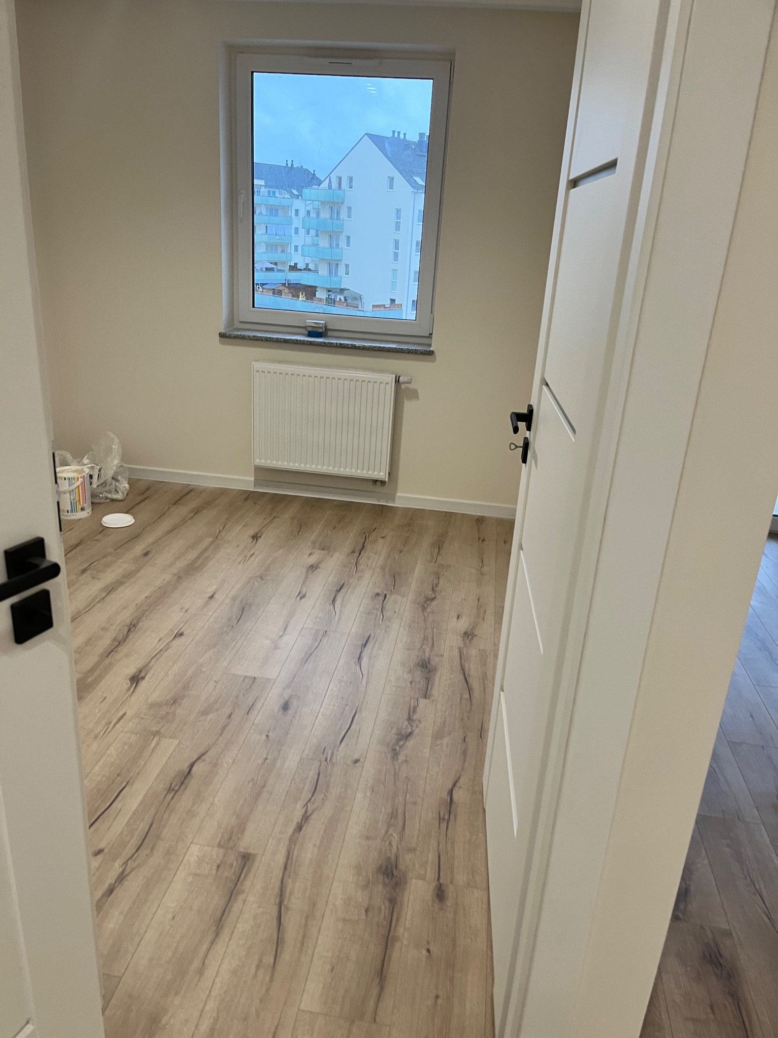 Pusty pokój po remoncie z panelami podłogowymi w odcieniach drewna, widoczne białe drzwi z czarną klamką, grzejnik pod oknem i wiadro z farbą w rogu.