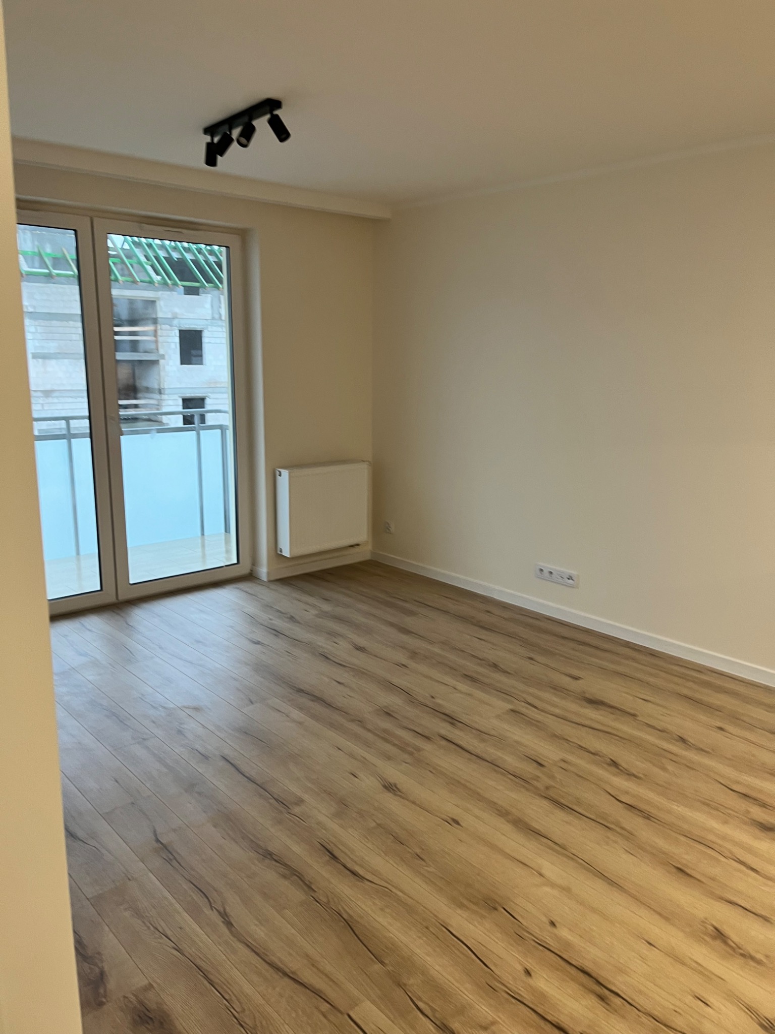 Pusty pokój z panelami podłogowymi w odcieniach brązu, białymi ścianami, grzejnikiem i oknem balkonowym z widokiem na budowę.