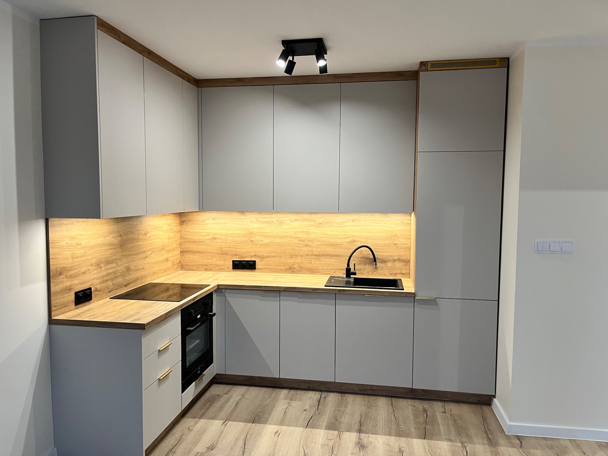 Nowoczesna kuchnia z szarymi szafkami, drewnianym blatem i oświetleniem LED pod szafkami górnymi. Czarny zlew i bateria, płyta indukcyjna. Widoczna wentylacja w górnej szafce.