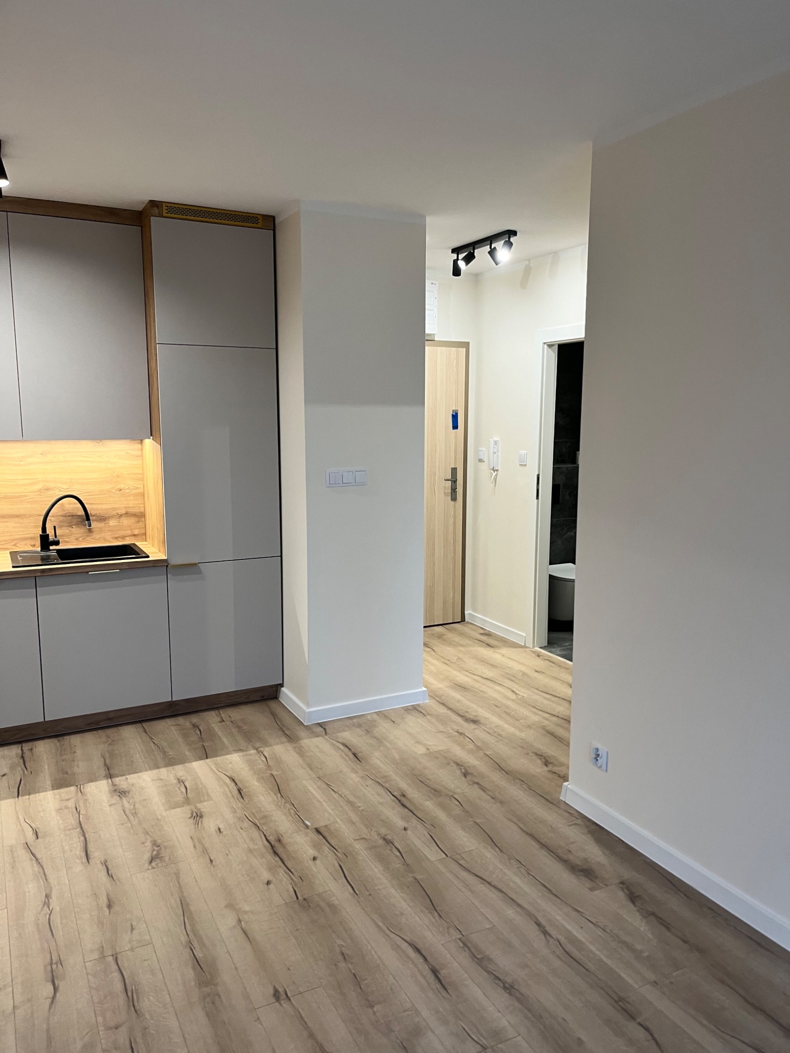 Nowoczesna kuchnia z szarymi szafkami, drewnianym blatem i czarnym zlewem, widok na przedpokój z drzwiami i łazienką w tle, podłoga laminowana z wzorem drewna.