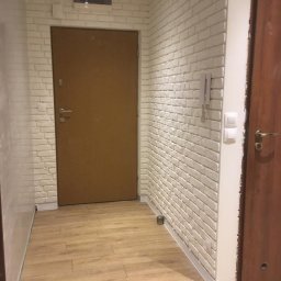 Mamut Company Sp. z o.o. - Korytarz po remoncie: ściany wyłożone białą cegłą dekoracyjną, podłoga z paneli imitujących drewno, dwoje drzwi w odcieniach brązu, widoczny domofon na ścianie.