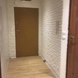 Korytarz po remoncie: ściany wyłożone białą cegłą dekoracyjną, podłoga z paneli imitujących drewno, dwoje drzwi w odcieniach brązu, widoczny domofon na ścianie.