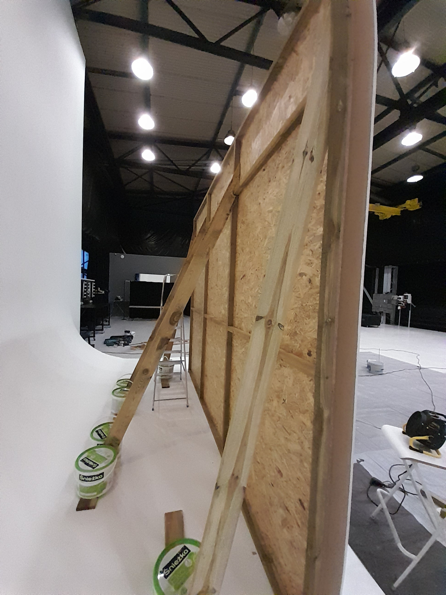 Konstrukcja z płyty OSB podparta drewnianymi belkami w studio, widoczne puszki z farbą Śnieżka i narzędzia na stole. Przygotowania do malowania ścian.