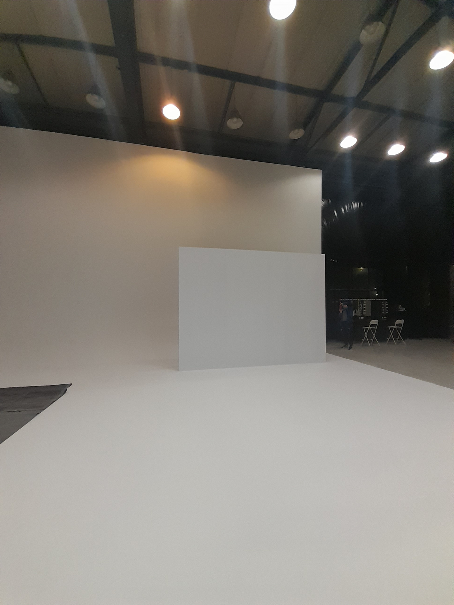 Pusta przestrzeń studyjna z białym tłem, oświetleniem sufitowym i osobą w tle, przygotowana do sesji zdjęciowej lub filmowej.