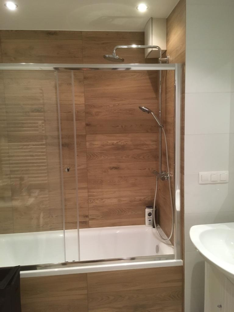 Biała wanna z szklanym parawanem, drewnopodobne płytki na ścianie, deszczownica i słuchawka prysznicowa, biała umywalka z szafką w rogu łazienki.