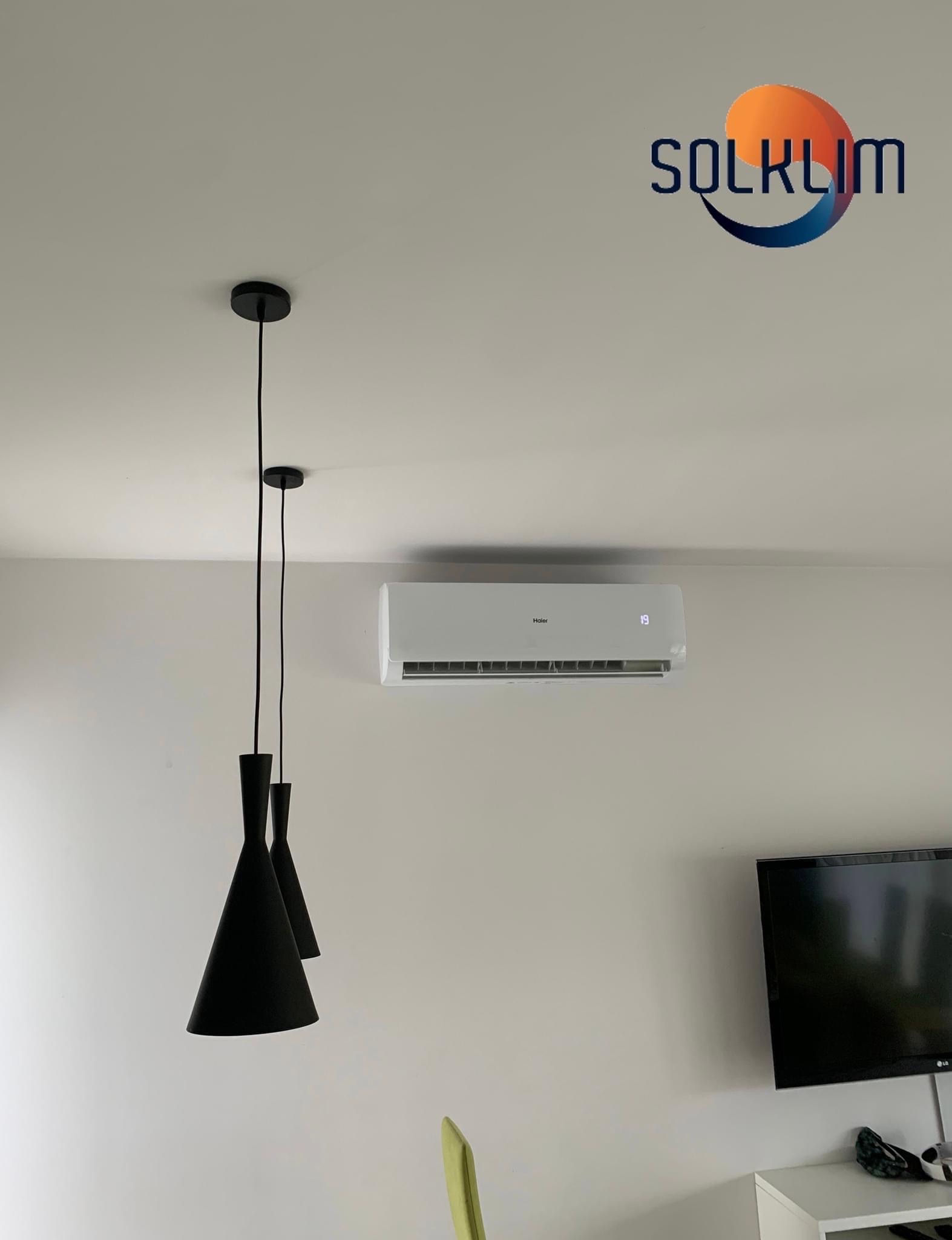 Biały klimatyzator Haier z wyświetlaczem temperatury 19 stopni, zamontowany na ścianie w salonie, widoczne czarne lampy wiszące i logo firmy Solklim.