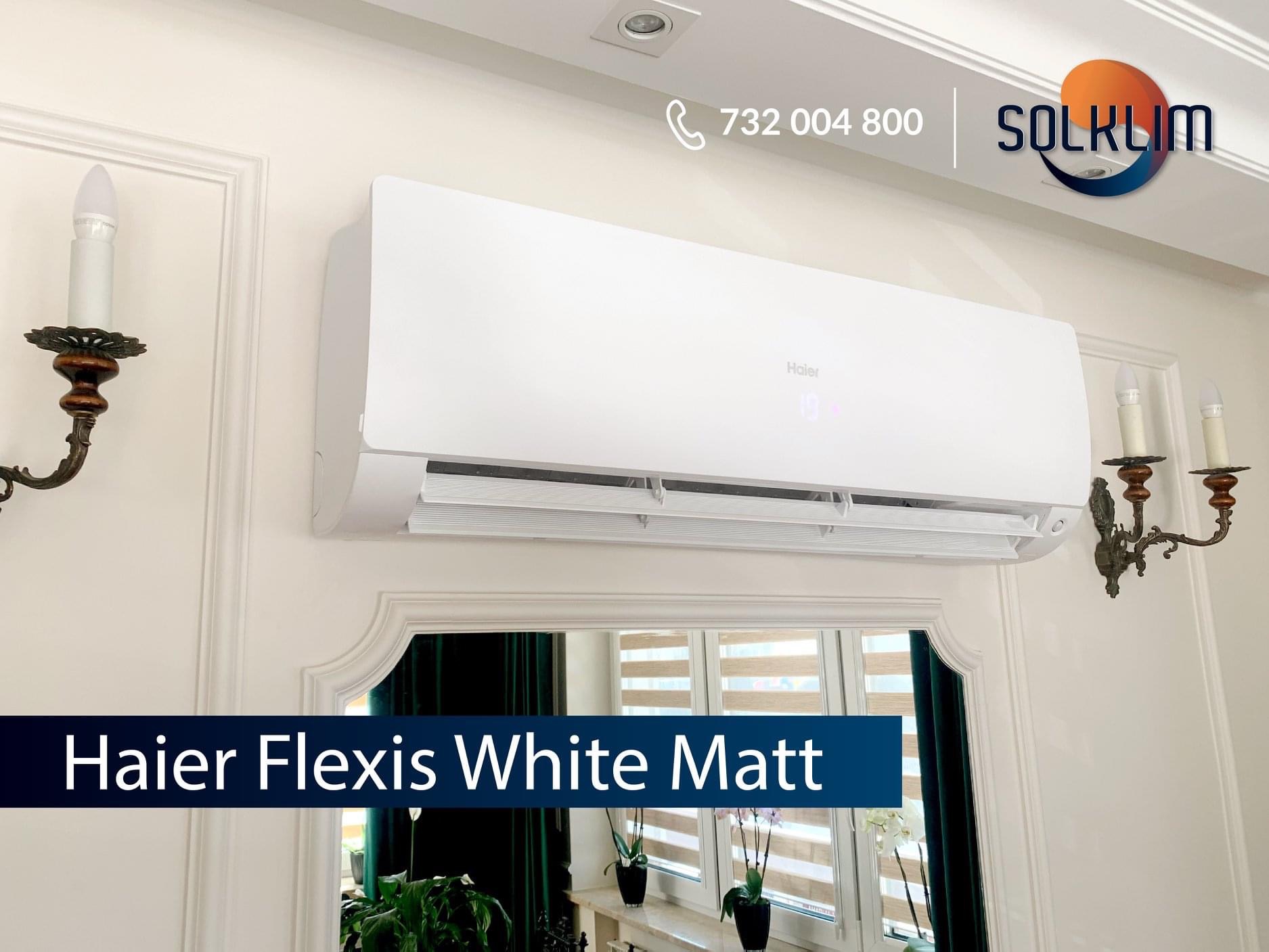 Biały klimatyzator Haier Flexis zamontowany na ścianie w pomieszczeniu z dekoracyjnymi kinkietami i lustrem. Widoczny numer telefonu firmy SOLKLIM.