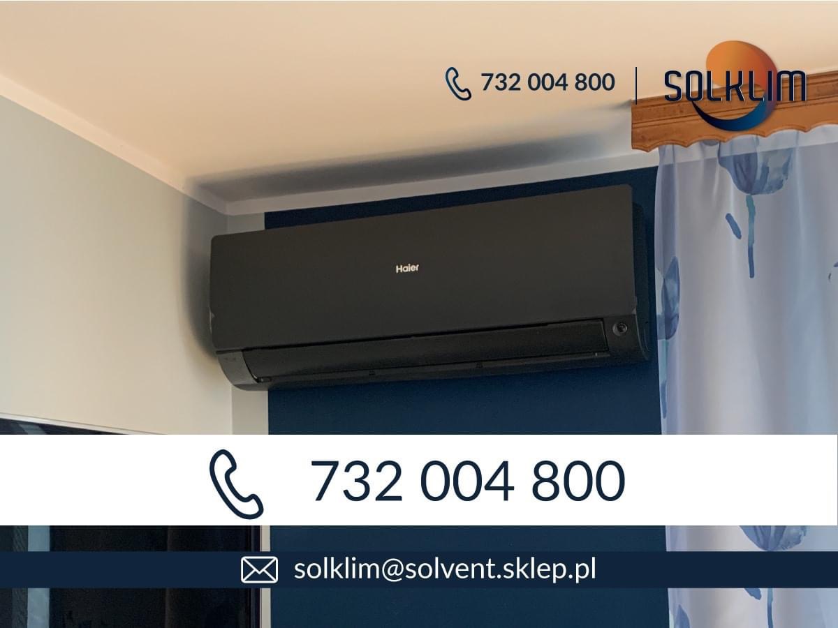 Czarny klimatyzator Haier zamontowany na ścianie pomalowanej w dwóch odcieniach niebieskiego, widoczne logo firmy i zasłona w tle.