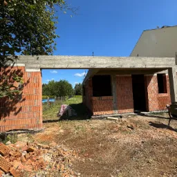 Budowa domu w stanie surowym otwartym, ściany z czerwonej cegły, widoczny betonowy strop, w tle drzewa i błękitne niebo.