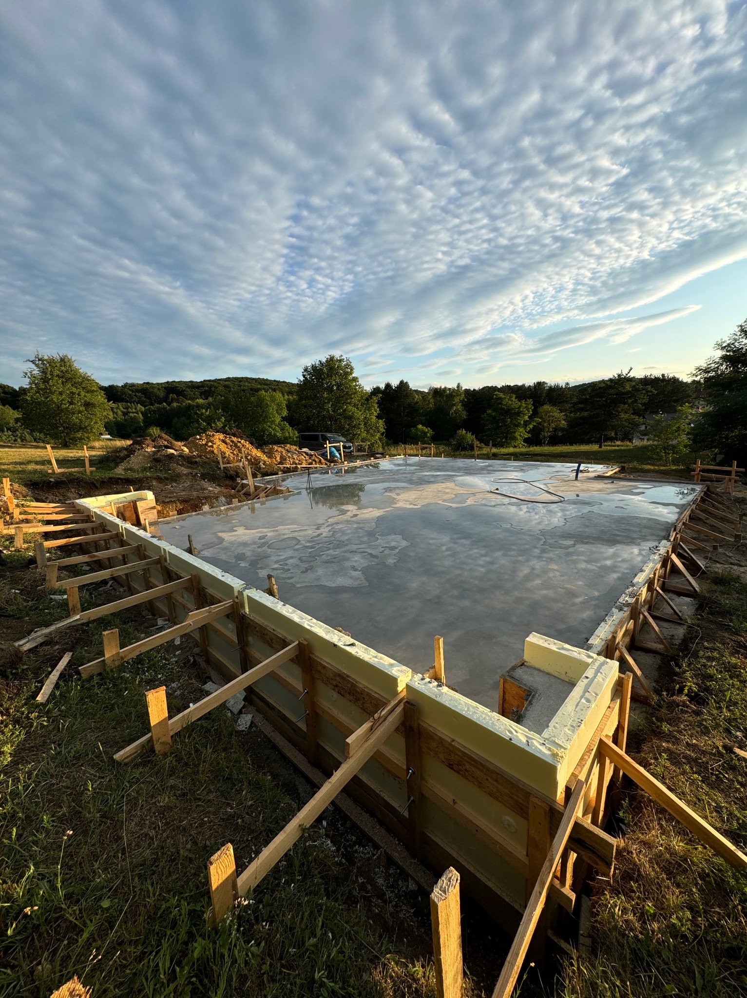 Wylany fundament płyty z drewnianym szalunkiem i izolacją termiczną z pianki, widoczne niebo z chmurami odbijające się w mokrym betonie.