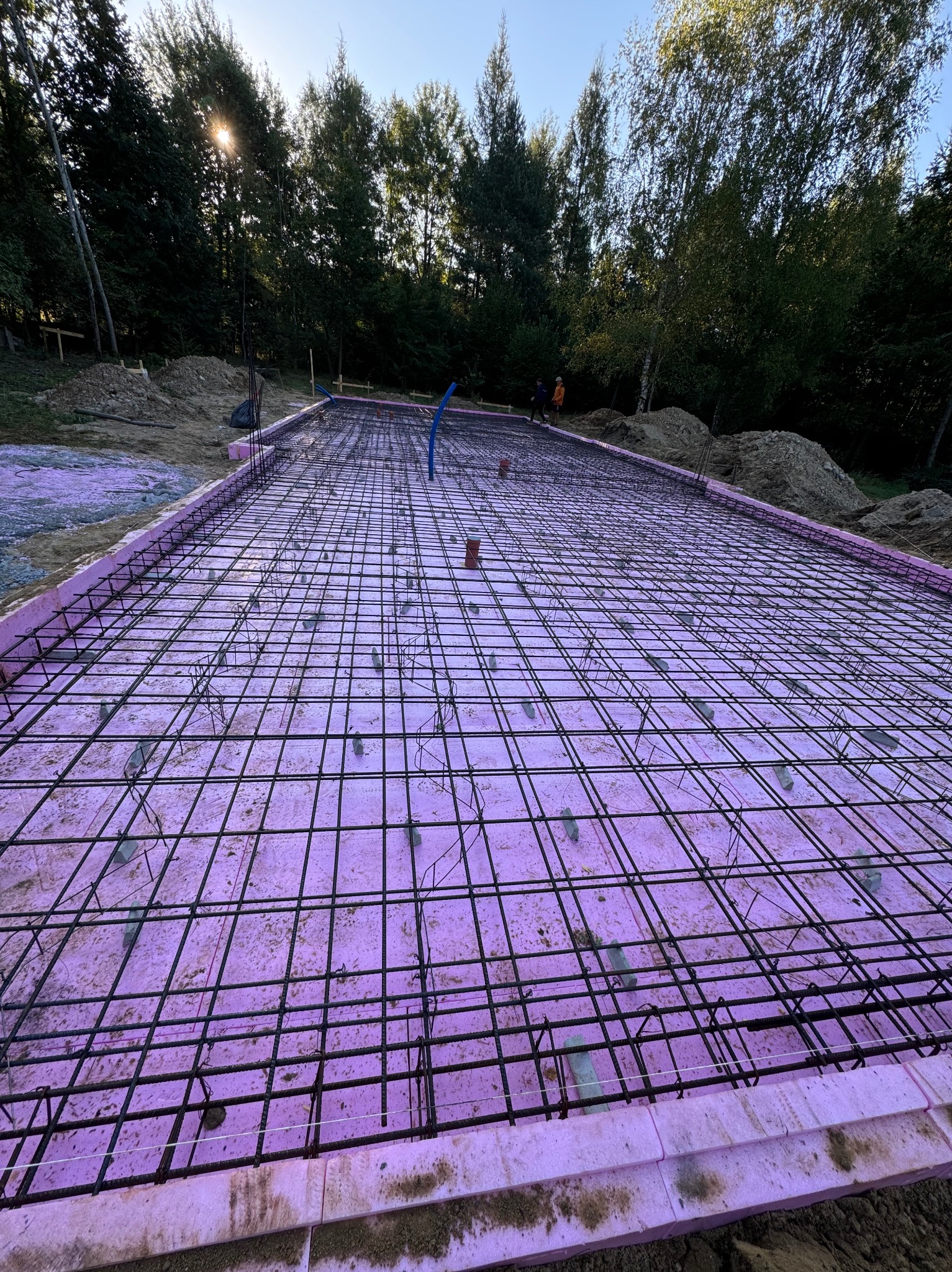 Przygotowanie płyty fundamentowej: różowy styropian, siatka zbrojeniowa z dystansami, widok z góry na tle drzew i nieba.