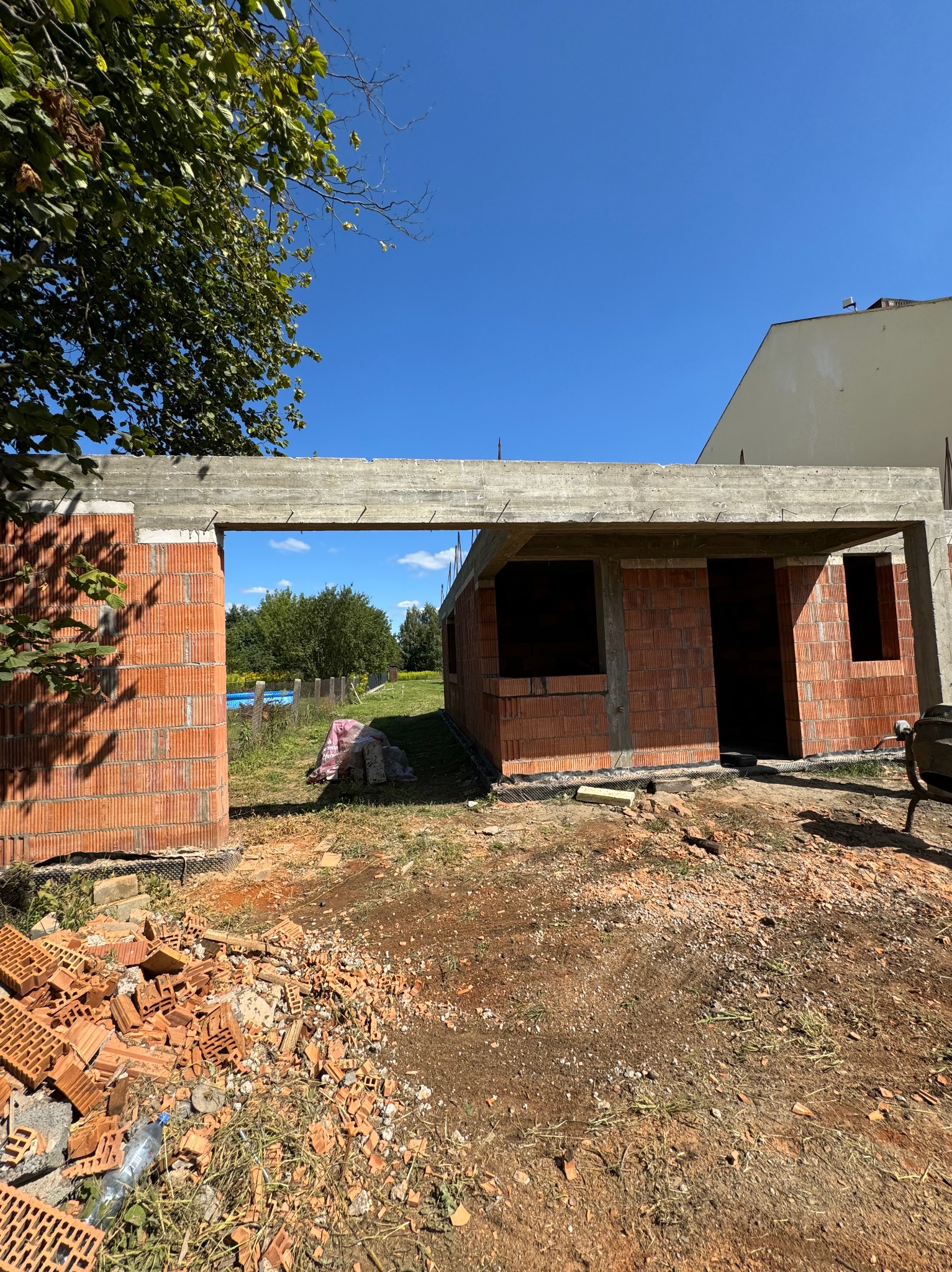 Budowa domu w stanie surowym otwartym, ściany z czerwonej cegły, widoczny betonowy strop, w tle drzewa i błękitne niebo.