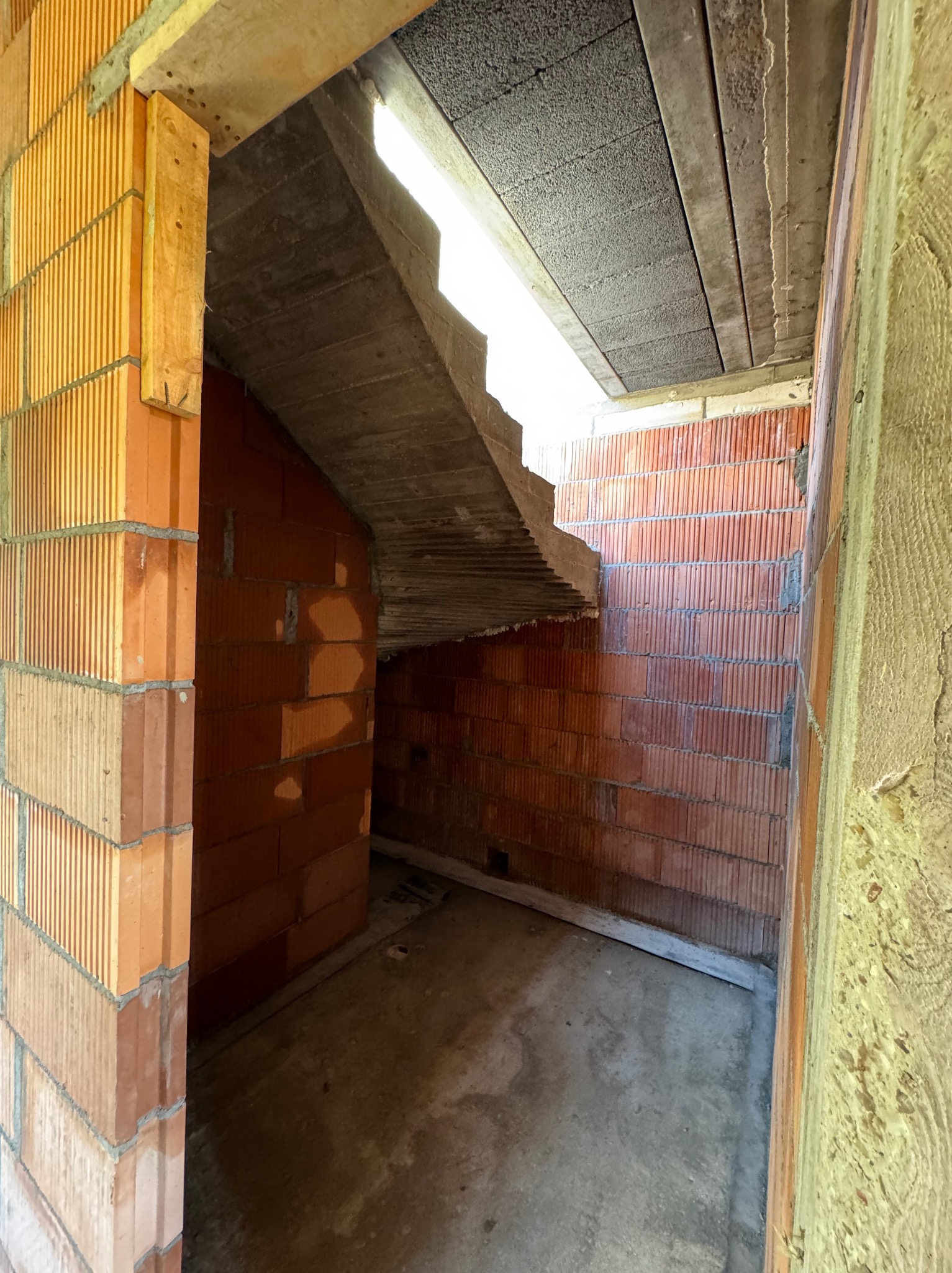 Surowy stan piwnicy z pustaków ceramicznych, widoczne betonowe schody i deski szalunkowe, w tle wpadające światło.