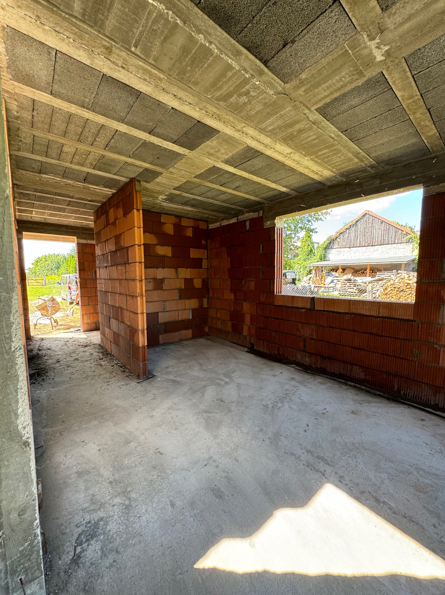 Wnętrze budynku w trakcie budowy: ściany z czerwonej cegły dziurawki, sufit z betonowych pustaków, widok na zewnątrz przez otwór okienny, w tle sterta drewna