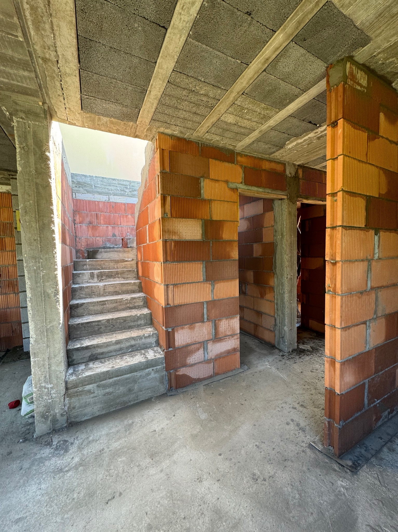 Wnętrze surowego budynku w trakcie budowy: widoczne betonowe schody, ściany z czerwonej cegły dziurawki, betonowy strop z pustaków szalunkowych, surowy beton na podłodze.