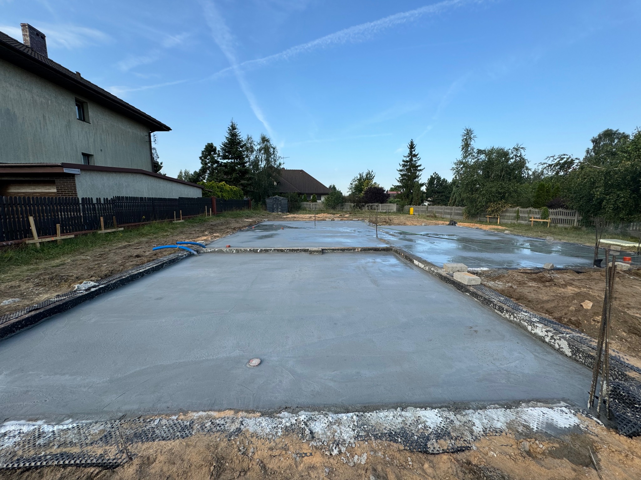 Wylany betonowy fundament pod budynek jednorodzinny, widoczne zbrojenia i izolacja pionowa, w tle dom i błękitne niebo z delikatnymi smugami kondensacyjnymi.