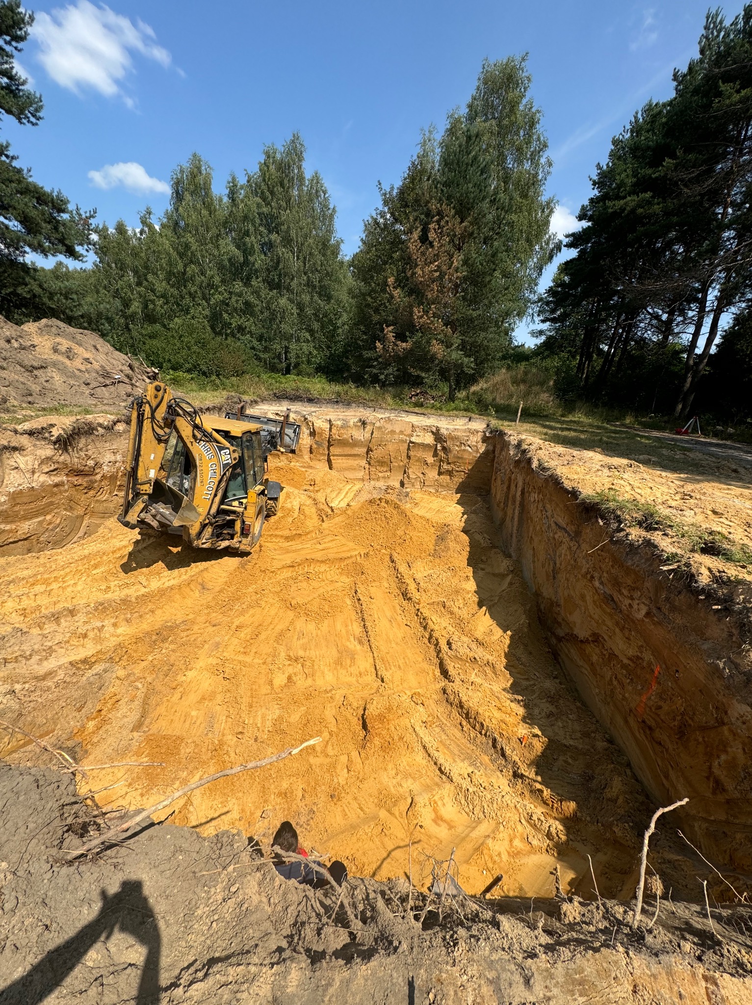 Żółta koparko-ładowarka CAT w wykopie pod budowę, widoczne warstwy ziemi, drzewa w tle, błękitne niebo