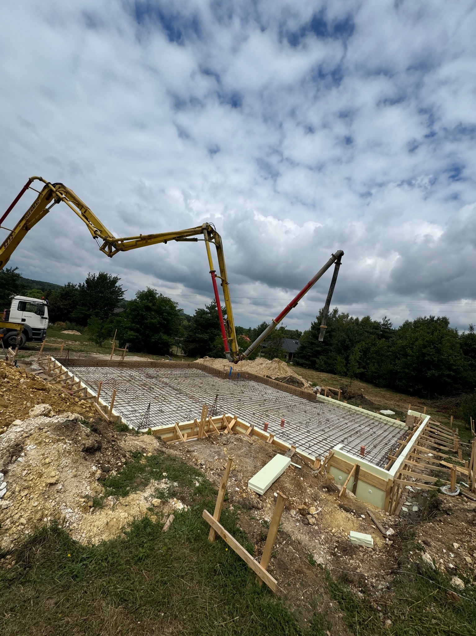 Wylewanie betonu na zbrojenie szalunku pod płytę fundamentową za pomocą pompy do betonu, widok z góry, pochmurne niebo w tle.