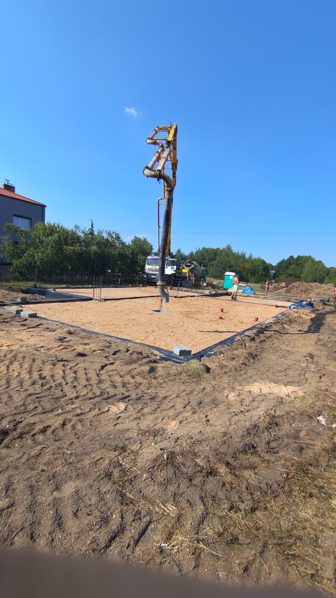 Wylewanie betonu za pomocą pompy do wykopu pod fundamenty, widoczne zbrojenia i przygotowany teren, w tle betoniarka i pracownicy.