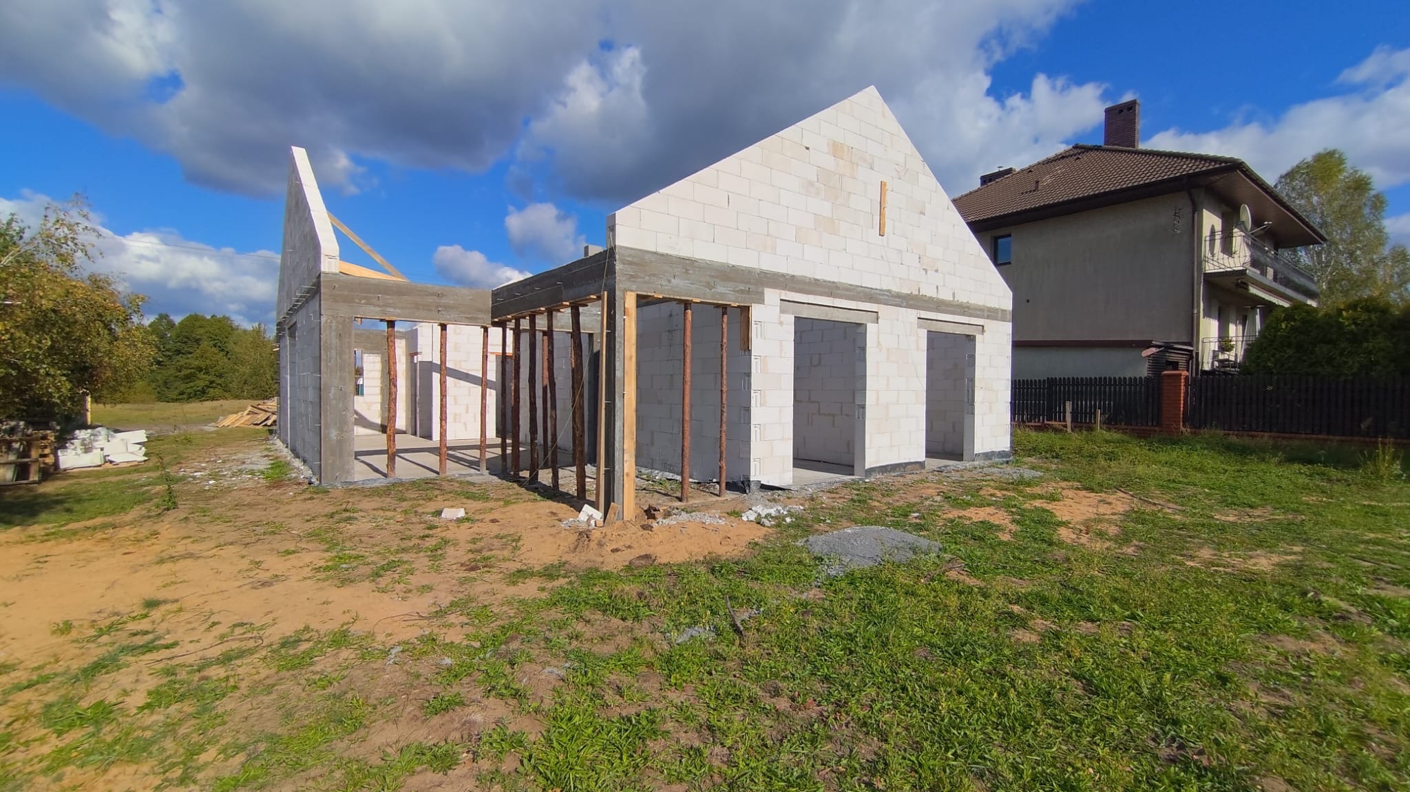 Budowa domu jednorodzinnego w trakcie wznoszenia ścian z bloczków betonowych, widoczne drewniane podpory i zbrojenia, w tle dom sąsiada.